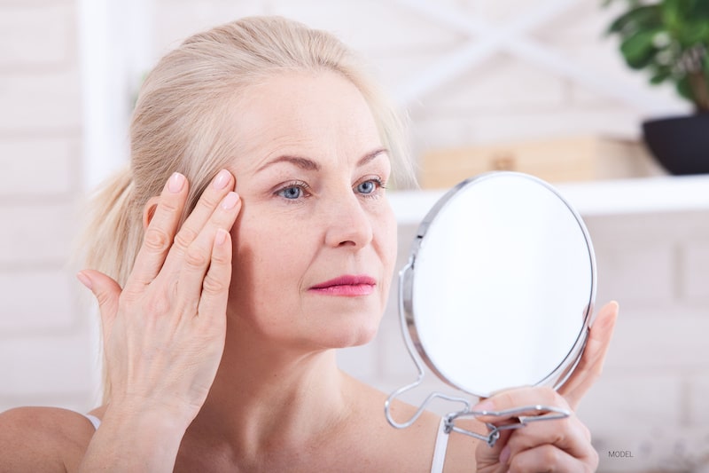 Middle-aged blond woman looking in a hand mirror while smoothing her facial skin