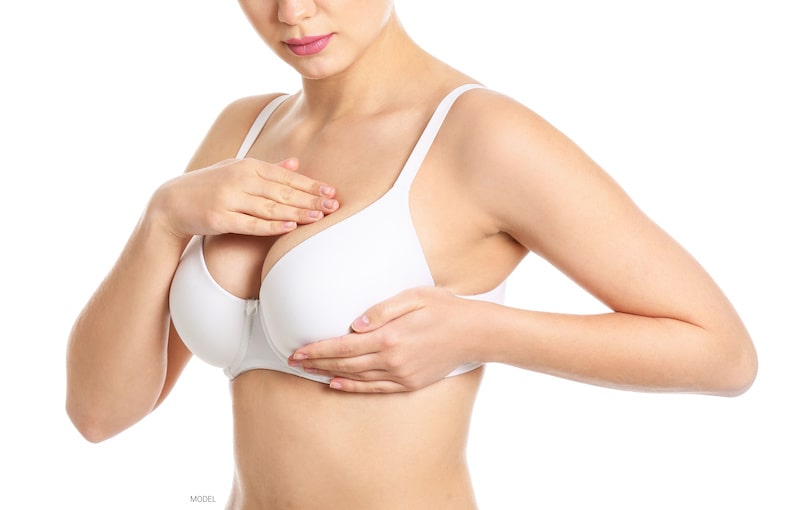 A woman in a white bra checks her breasts.