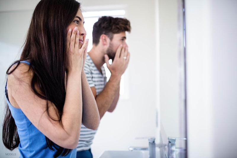 Attractive woman and man looking at their reflection in the mirror while touching their faces